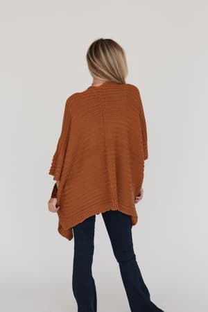 A woman with long blonde hair faces away, wearing the So Deserving Knit Ruana in camel with dark flared jeans—a chic, boho-inspired look—against a plain light background.