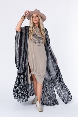 A woman poses against a white background wearing a wide-brim tan hat, a tan Free Bird graphic dress with a front knot, the Paisley Tapestry Free Flow Duster Kimono in black, cream ankle boots, and multiple bracelets.