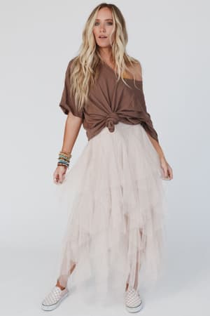 A woman stands against a white background, wearing a loose brown t-shirt knotted at the waist, the Boho Ballerina Skirt - Nude with layered tulle, white slip-on shoes, and colorful bracelets on her left wrist.