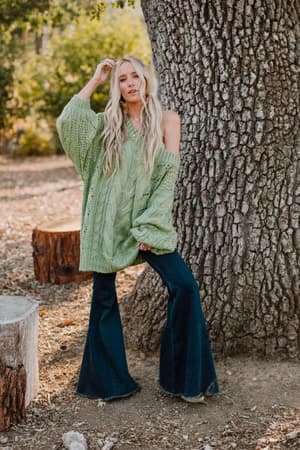 A woman with long blonde hair stands outdoors, leaning against a tree while wearing the Golden Hour Bubble Sleeve Sweater Dress in light green.