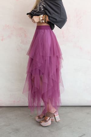 A person stands sideways wearing the Boho Ballerina Skirt - Mauve in layered tulle, a black embroidered top, multiple bracelets, and colorful platform heels against a light wall with pink accents.