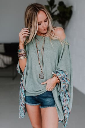 A woman with long blonde hair wears the Fallon Embroidered Sleeve Poncho in sage, paired with denim shorts, stacked bracelets, and layered necklaces. She stands indoors, looking down with one hand in her hair.