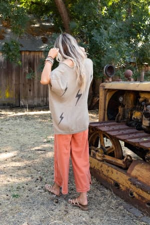 A woman stands outdoors with her back turned, adjusting her hair. She wears the Struck By You Dolman Sleeve Sweater Top in Mocha with black lightning bolt designs, coral pants, and sandals near rusty construction equipment and trees.