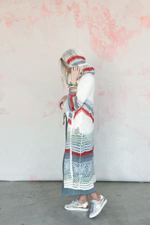 Wearing the Love In Layers Cardigan - Multi, a person stands sideways by a pink and white wall. The long, hooded cardigan features red, gray, and white stripes for a relaxed bohemian look with casual shoes and bracelets.
