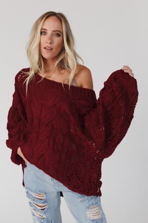 A woman with long blonde hair wears the Golden Hour Bubble Sleeve Sweater Dress in burgundy, featuring oversized bubble sleeves and an off-the-shoulder design, standing against a plain light background.