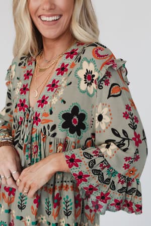 A smiling woman wears the Leona Floral Print Tunic in sage, featuring a light green floral print, ruffled sleeves, and a v-neckline. She accessorizes with layered gold necklaces and rings on her fingers.