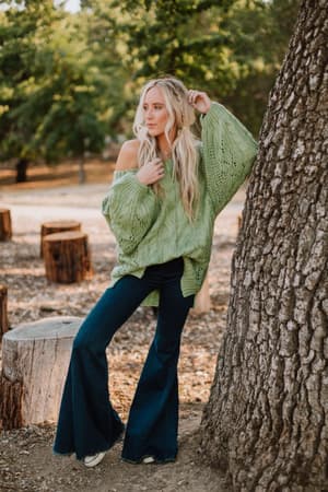 A woman with long blonde hair leans against a tree outdoors, wearing the Golden Hour Bubble Sleeve Sweater Dress in light green. Tree stumps and greenery are visible in the background.