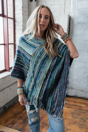 A woman with long blonde hair wears the Madeleine Poncho - Blue, featuring colorful stripes and fringe, paired with ripped jeans. She stands in a rustic room with white brick walls, wooden floors, and stacked bracelets on both wrists.