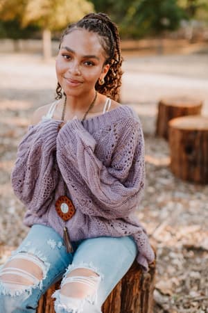 A woman with braided hair sits on a tree stump outdoors, smiling gently and holding her hands to her chest, wearing the Golden Hour Bubble Sleeve Sweater Dress in Lavender. Trees and more stumps fill the background.