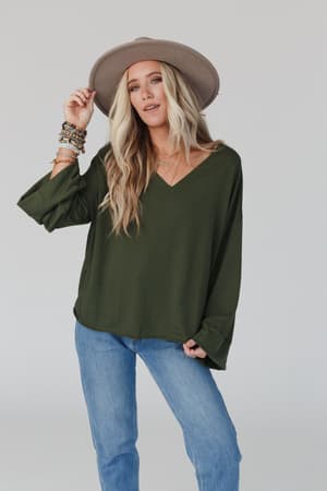 A woman in The Nest Love It V Neckline Sweater Top - Olive, blue jeans, and a wide-brimmed hat poses against a plain light background. She has long wavy blonde hair and holds her hat's brim with one hand.