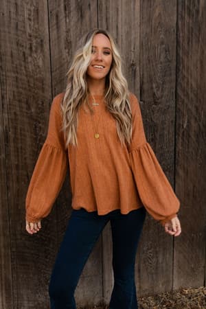 A woman with long, wavy blonde hair smiles in front of a wooden wall, wearing The Nest Sweet Heart Balloon Sleeve Knit Sweater in Burnt Orange, paired with dark jeans and gold necklaces.