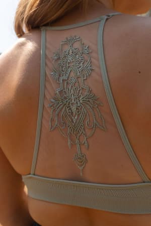 Close-up of a woman wearing the Tattoo Bralette - Sage, a light gray mesh racerback top with intricate floral embroidery down the center. Her light brown hair falls over her shoulder, highlighting the delicate, tattoo-inspired design.