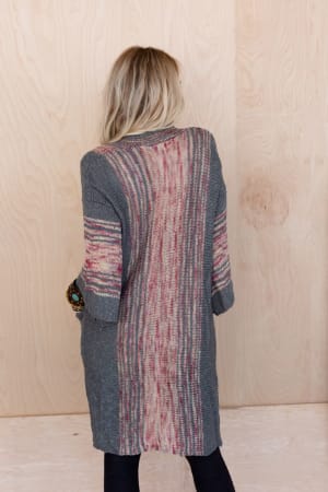 A woman with blonde hair faces a light wooden wall, wearing the Rose Boho Throw On Cardigan in gray featuring multicolored vertical stripes and oversized pockets, paired with dark pants.
