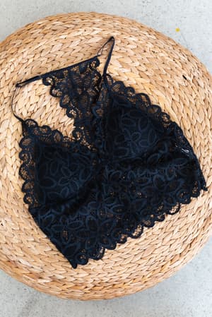 The Eye Of The Sun Padded Bralette - Black, featuring intricate floral crochet lace, is laid flat on a round woven straw mat against a light gray background.