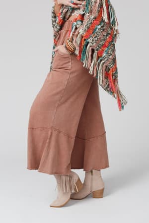 A person wearing the Feeling Good Wide Leg Pant in Faded Rust, beige fringe ankle boots, and a colorful fringed shawl displays chic boho style; only their lower half is shown against a plain grey background.