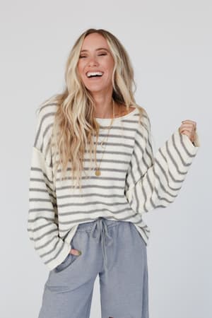 A smiling woman with long blonde hair wears the All My Stripes Oversized Sweater in Ivory Gray and loose gray pants, accessorized with a gold necklace. She stands against a simple light background, one hand in her pocket.