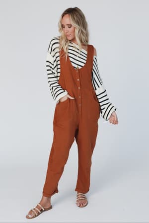 A woman with wavy blonde hair stands against a plain background, gazing to the side in a rust jumpsuit layered over the All My Stripes Oversized Sweater in cream and black, one hand casually slipped into her pocket.