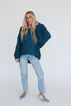 A woman with long blonde hair wears the Golden Hour Bubble Sleeve Sweater Dress in teal, paired with ripped light blue jeans and checkerboard slip-on shoes, standing against a plain white background.