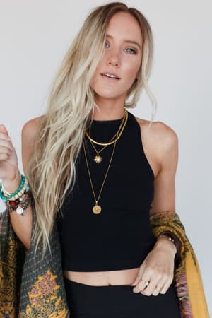 A woman with long, wavy blonde hair wears a black High Neck Racerback Brami, layered gold necklaces, several bracelets, and a patterned shawl over one arm as she stands against a plain light background.
