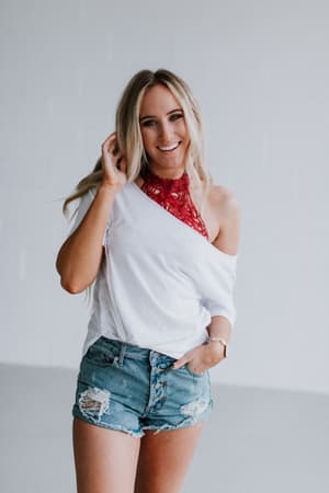 A smiling woman with long blonde hair wears a white off-the-shoulder top, distressed denim shorts, and the Baroque High Neck Velvet Bralette in Wine. She stands in a bright, minimal space, touching her hair with one hand.