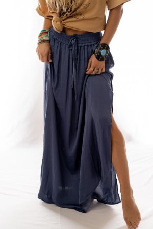 A person stands barefoot against a white background, wearing a tan knotted t-shirt and the Stasia Maxi Skirt - Blue, which is high waisted, flowy, has a high side slit, and is accessorized with bracelets and a large cuff.
