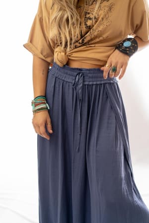 A person wearing a knotted tan graphic t-shirt and the Stasia Maxi Skirt - Blue, accessorized with multiple bracelets, rings, and a wide black wrist cuff. Their face is not visible.