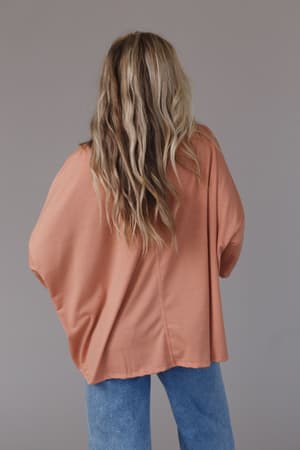 A person with long, wavy, blonde hair stands facing away, wearing The Nest Cozy Cool Oversized Tee in Coral and blue jeans against a plain gray background.
