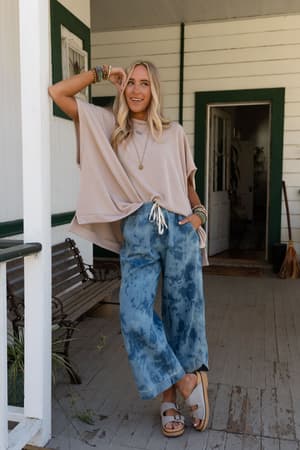 A woman with long blonde hair stands on a porch, smiling, in a loose beige top and The Nest So Comfy Wide Leg Denim Pant - Bleach Dye Denim, paired with sandals and layered necklaces, resting one arm on the doorframe.