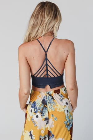 A woman with long blonde hair is seen from the back wearing the Light Of The Moon Crochet Trim Bralette in blue gray, featuring multiple thin straps in a striking geometric pattern.