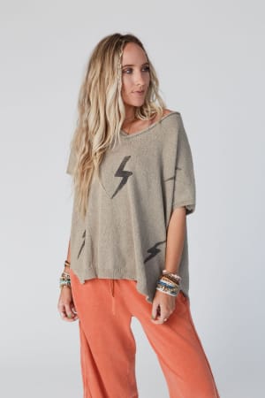 A woman with long, wavy blonde hair wears the Struck By You Dolman Sleeve Sweater Top in mocha, featuring black lightning bolt designs. She pairs its loose silhouette with coral pants and beaded bracelets against a plain light background.