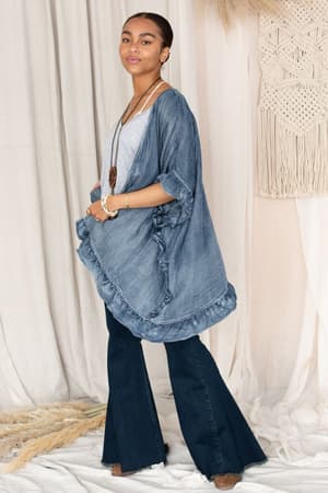 A woman wearing the Robbie Denim Jacket - Blue stands confidently in front of cream curtains and woven wall decor.