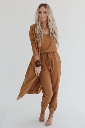 Barefoot blonde woman stands against a white background wearing the Set to Go Textured Jumpsuit And Cardigan Set in camel, accessorized with jewelry and a chunky bracelet.