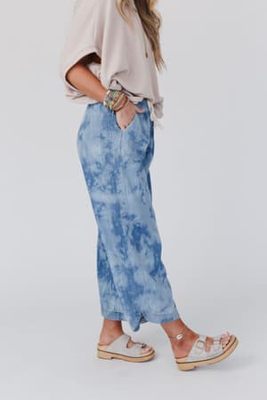 A woman stands in a relaxed pose, wearing The Nest So Comfy Wide Leg Denim Pant - Bleach Dye Denim with a beige oversized top and sandals. She has her hands in her pockets, loose hair, multiple bracelets, and the background is white.