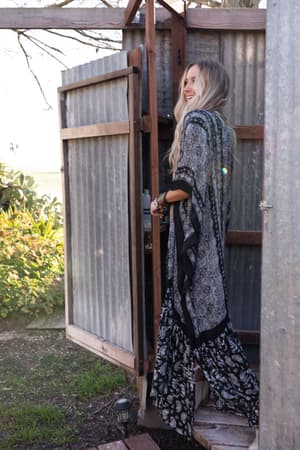 A woman with long blonde hair stands outside in the sunlight, wearing the Paisley Tapestry Free Flow Duster Kimono - Black, holding the door of a rustic corrugated metal shed with greenery in the background.
