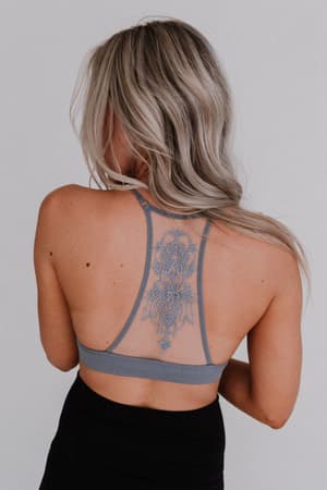 A woman with long blonde hair faces away from the camera, wearing the Tattoo Bralette - Gray with black bottoms, revealing an intricate blue tattoo running down the center of her upper back.