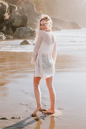 A woman stands barefoot on the beach, wearing the Brenden Knit Netted Cardigan in Ivory as a sheer cover-up. She glances over her shoulder with windblown hair, sunlight, waves, and rocks in the background.