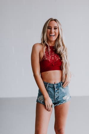 A smiling woman with long blonde hair wears the Baroque High Neck Velvet Bralette in wine and ripped denim shorts, standing against a plain white background.