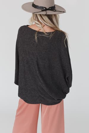 A woman with long blonde hair, wearing a taupe wide-brimmed hat and The Nest Fly With Me Batwing Sleeve Knit Sweater in Charcoal, stands with her back to the camera against a light background.