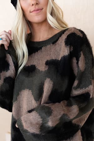 A woman with long blonde hair models the Wild Wanderer Sweater - Camo, a cozy, loose-fit camouflage-patterned sweater in black, brown, and green, standing against a light wooden background. Her face is partially visible.