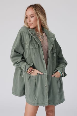A woman with long blonde hair and layered necklaces stands looking down to the side with her hands in the pockets of the Layers On Layers Perfect Boho Parka in olive, a loose fit jacket featuring large front pockets and snap buttons.
