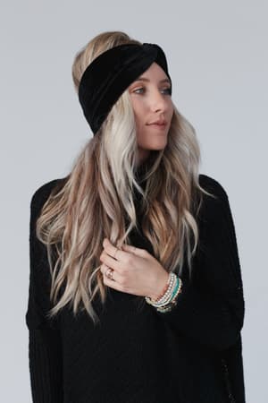 A woman with long, wavy blonde hair wears the Everyday Velvet Headband in Black and a black knit sweater. She has several beaded bracelets on her wrist and gazes slightly to the side against a plain light background.