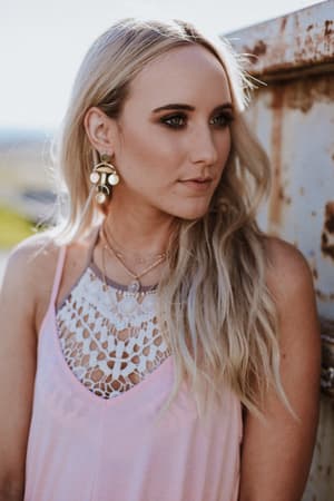 A woman with long blonde hair wears the Crochet High Neck Bralette in Mocha, styled with layered necklaces and large earrings. She stands outdoors by a rusted metal structure, gazing to the side with a calm expression.
