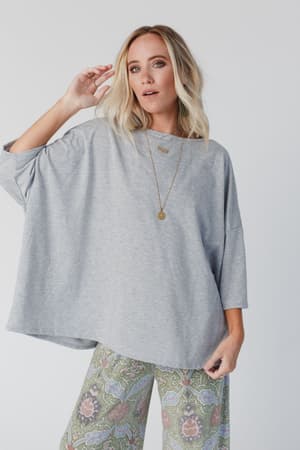 A blonde woman wearing The Nest Cozy Cool Oversized Tee in Heather Gray, paired with patterned wide-leg pants and a long gold necklace, stands against a light background, one hand near her face as she looks off to the side.