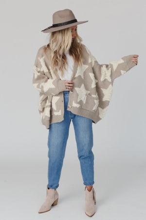 A woman wears the Wrapped In Stars Oversized Cardigan - Taupe, a chunky beige knit with bold white stars, paired with light blue jeans, a beige hat, and ankle boots. She stands against a plain background, gazing to the side with one arm raised.