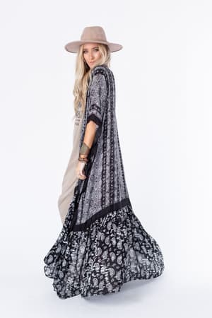 A woman in the Paisley Tapestry Free Flow Duster Kimono - Black stands against a white background, wearing a wide-brimmed hat and graphic tee, looking over her shoulder.