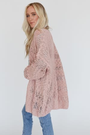 A woman with long blonde hair models the Cara Oversized Cardigan in pink with light blue jeans, standing sideways and looking at the camera against a plain white background.