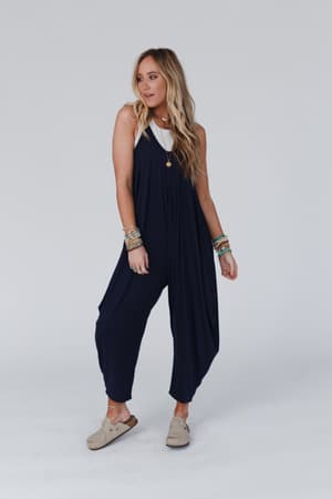 A woman with long blonde hair wears The Perfect Harem Jumpsuit in navy, paired with beige slip-on shoes, layered necklaces, and multiple bracelets, standing against a plain light background.