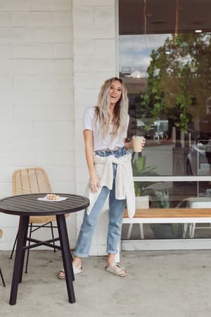 A woman with long, wavy blonde hair smiles outside a cafe, holding an iced drink. She wears the Scoop Neck Bralette Tee in white with blue jeans and sandals. A light sweater is tied at her waist; a pastry sits on a small table nearby.