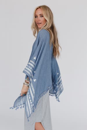 A woman with long blonde hair models the Fallon Embroidered Sleeve Tassel Poncho in blue, featuring geometric white embroidery and tassels, layered over a light gray dress against a plain white background.