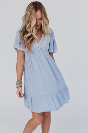 A woman with long, wavy blonde hair wears the Magdaline Mini Dress - Blue, featuring short sleeves and a knee-length hem, standing against a plain background and gazing downward to the side.